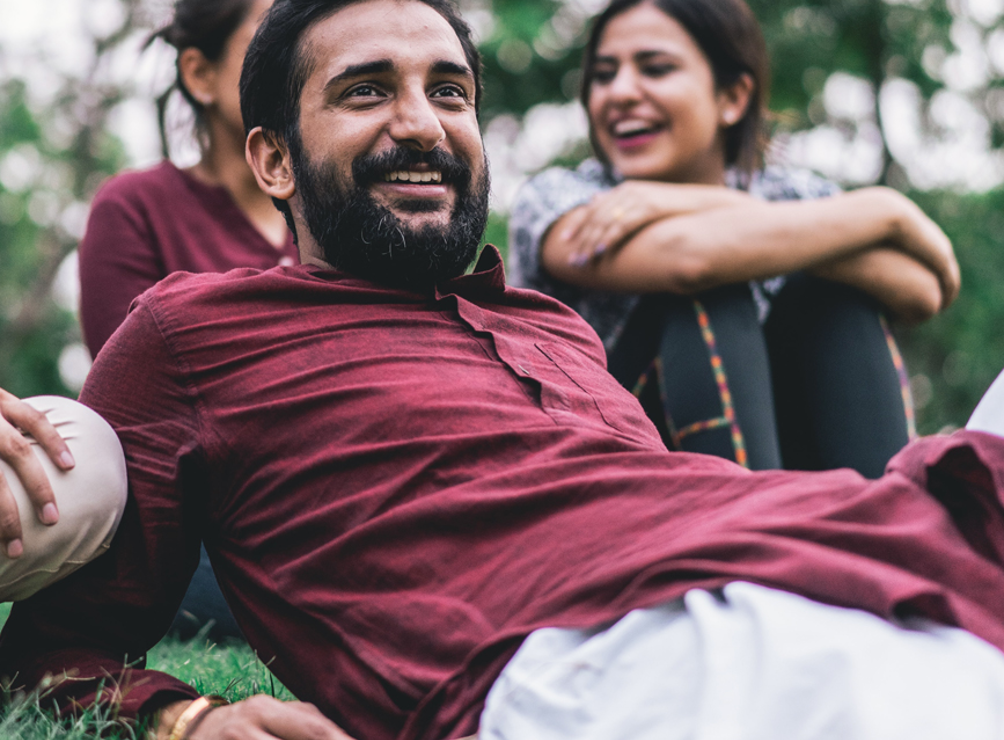 Man laying on the grass outside. He and other around him are smiling. 