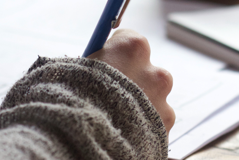 Close up of a hand writing on white paper on a table. They are writing with a blue pen.