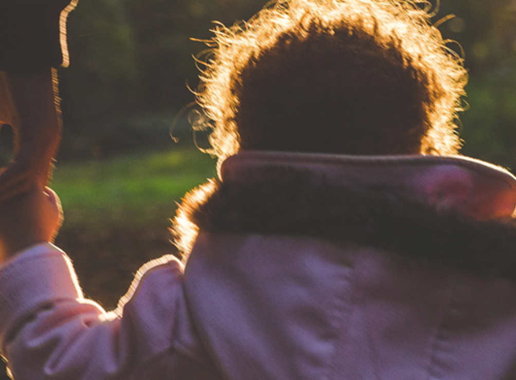 Young child walking with her back to us. She is holding the hand of an adult. They're walking in the sun.